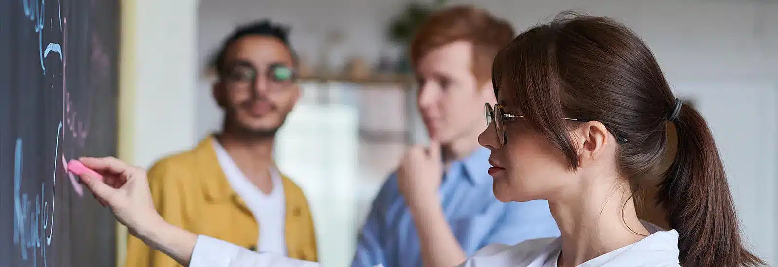 Frau mit langen dunklen Haaren schreibt an die Tafel. Zwei Herren schauen im Hintergrund zu.