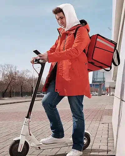 Mann in orangener langer Jacke auf einem E-Roller mit Handy in der Hand.
